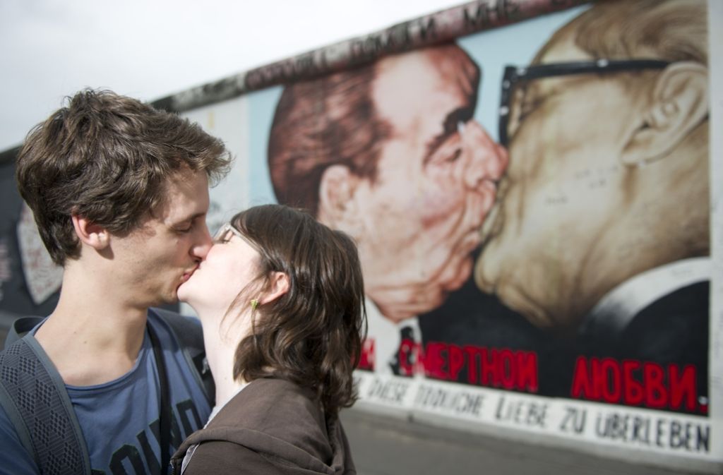 Zwei Touristen küssen sich in Berlin an der East Side Gallery vor dem Bild „Bruderkuss“. Der Bruderkuss zwischen Erich Honecker und Leonid Breschnew von Künstler Dmitri Wrubel gehört zu den bekanntesten Motiven der East Side Gallery.