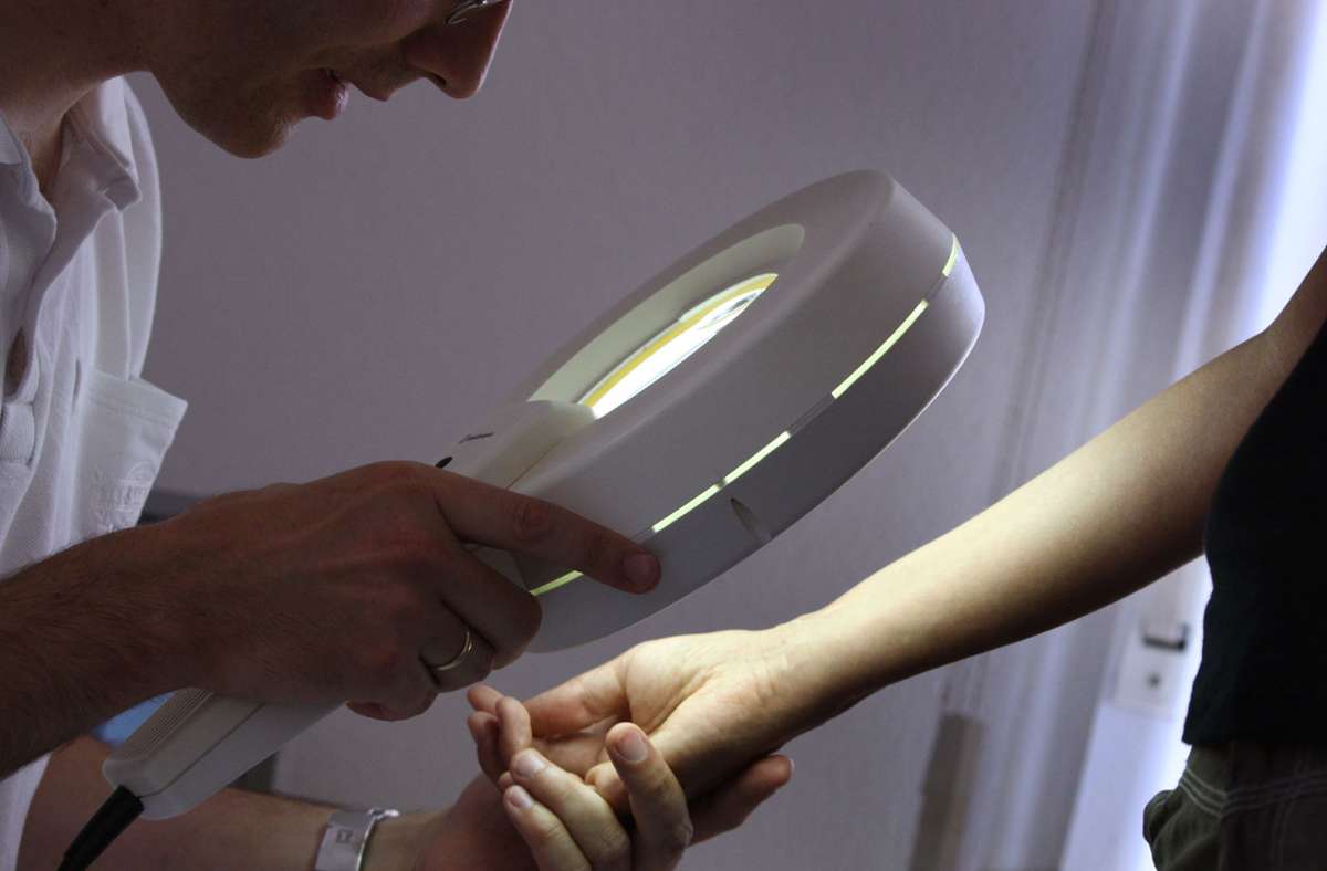 Ein Hautarzt untersucht in seiner Praxis mit einem Vergrößerungsglas die Haut einer Patientin bei einer Hautkrebs-Früherkennung.